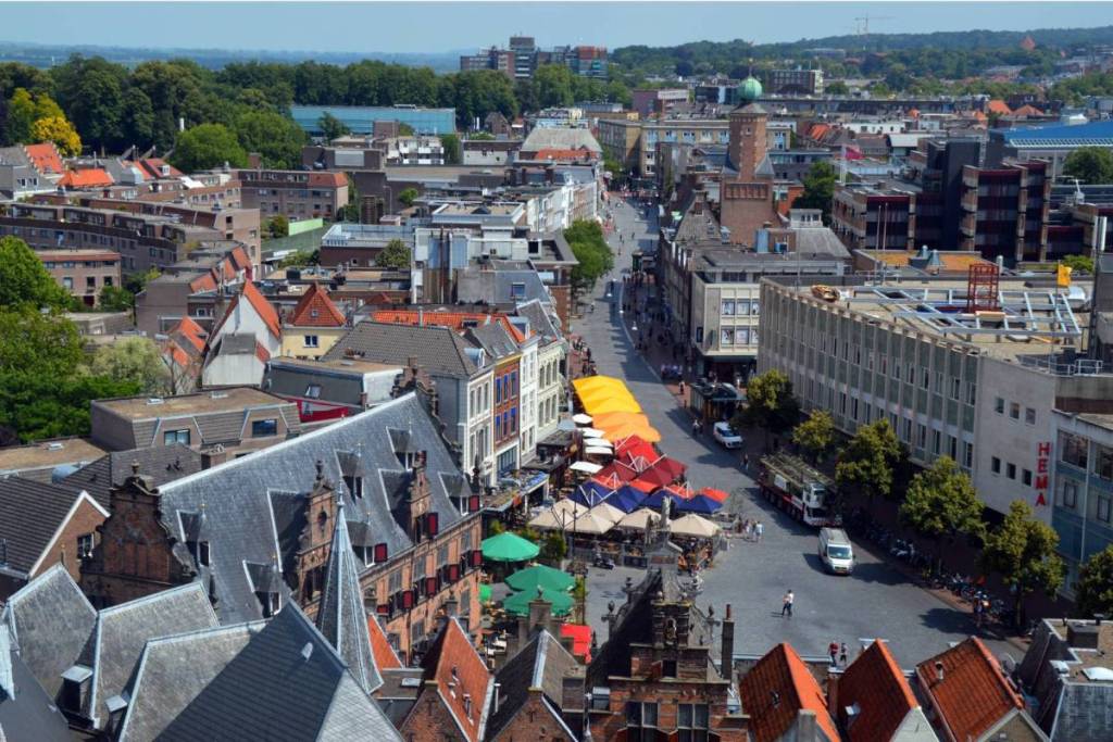 A panoramic bird’s-eye view of the historic Grote Markt and the rooftops of Nijmegen’s city center, with outdoor cafes, colorful umbrellas, and shoppers, as seen from the Stevenskerk.