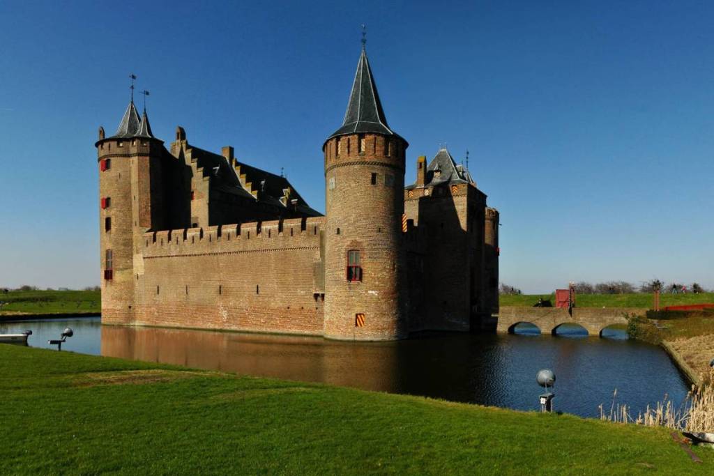 Une vue latérale ensoleillée du château médiéval de Muiderslot, entouré d’un fossé paisible. La forteresse en briques présente ses créneaux caractéristiques, ses tours d’angle pointues aux volets rouges et un pont en arc en pierre sur la droite, photographiée depuis une pente herbeuse sous un ciel d’un bleu limpide.