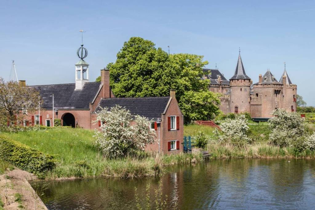 Une vue pittoresque sur le château de Muiderslot depuis l’autre rive, par une journée ensoleillée. Au premier plan, on aperçoit des dépendances historiques en briques aux volets rouges, entourées d’arbres verdoyants, d’arbustes blancs en fleurs et d’une pente herbeuse le long des douves.