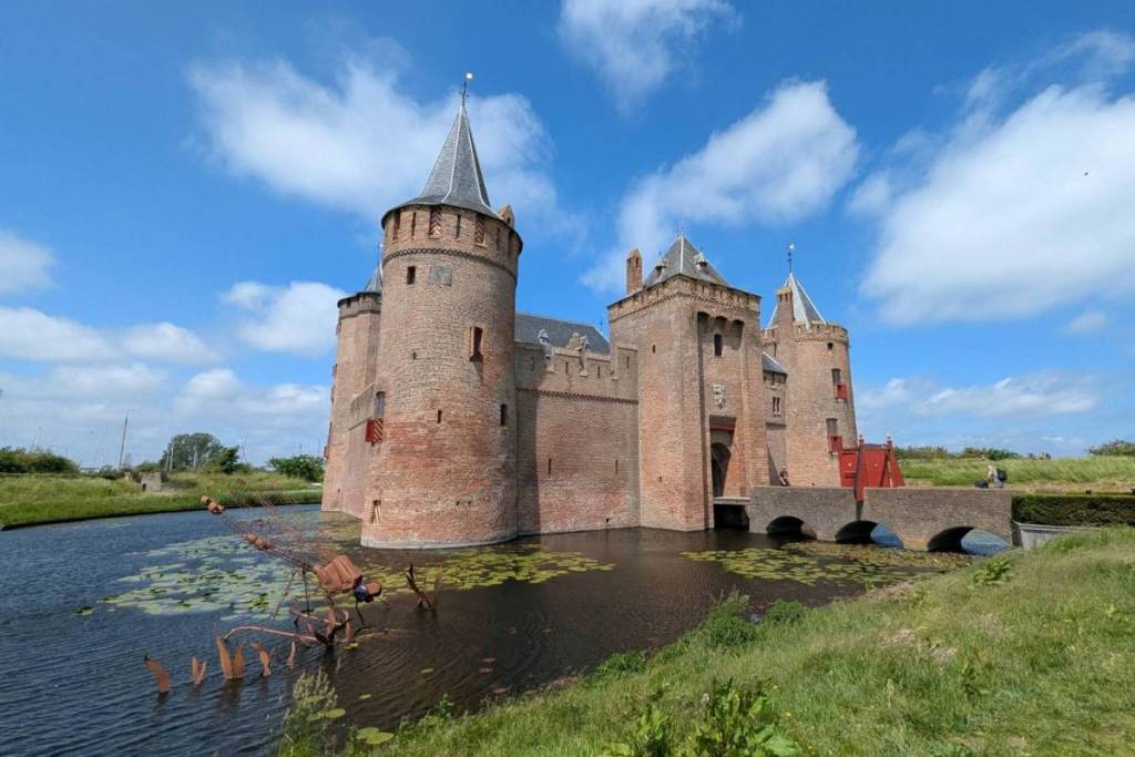 Une vue d'ensemble du château médiéval de Muiderslot à Muiden, sous un ciel bleu parsemé de nuages. Le château est entouré d'un fossé orné de nénuphars et dispose d'un pont en arc en pierre qui mène à l'entrée.