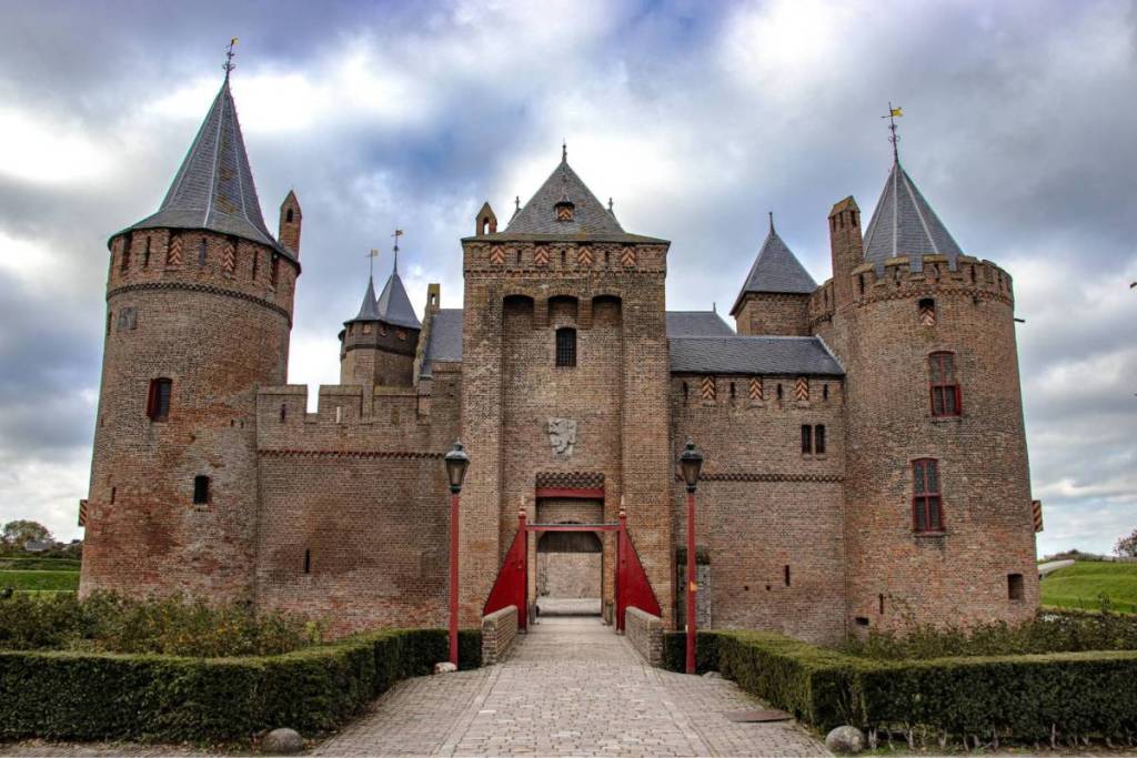 Vue de face de l’imposante porte d’entrée médiévale et du pont-levis du Muiderslot. Le château en pierre présente des tours d’angle robustes, des créneaux et des détails rouges, photographié depuis l’allée d’accès sous un ciel nuageux spectaculaire.