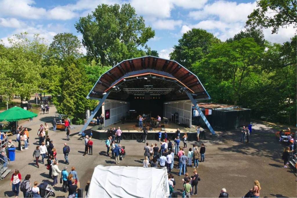 Luchtfoto van het openluchttheater in het Vondelpark, met een optreden op het grote boogpodium en een publiek dat zich verzamelt op het plein omringd door groene bomen.