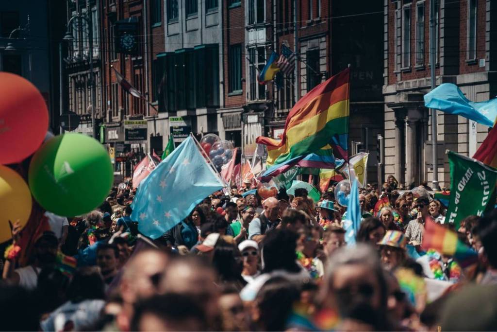 Een kleurrijke en drukke Pride-parade in een stadsstraat met historische gebouwen, waar een menigte zwaait met regenboogvlaggen en grote ballonnen.