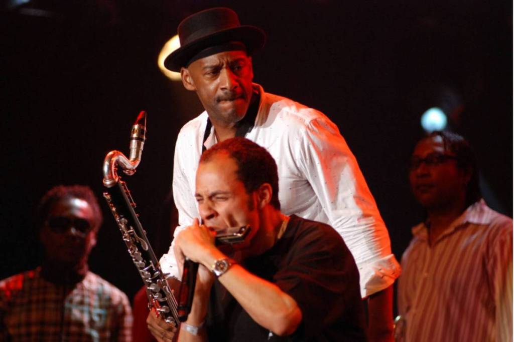 Live-optreden tijdens een jazzfestival in Amsterdam met een muzikant die intensief op een mondharmonica speelt, geflankeerd door een saxofonist met een hoed op een sfeervol verlicht podium.