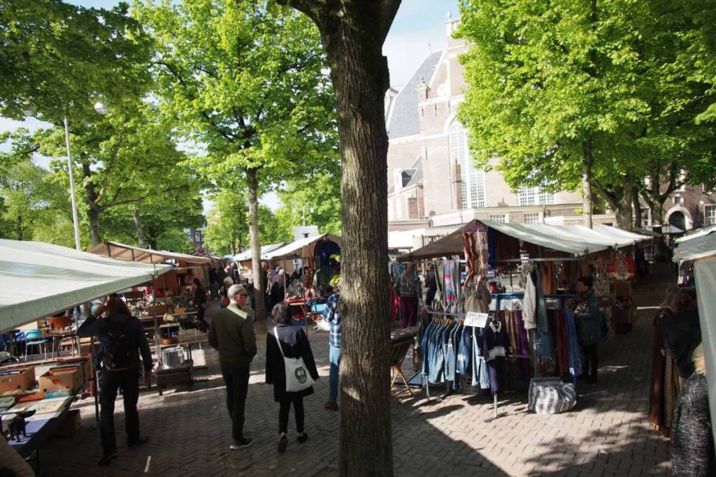 Bezoekers struinen langs kledingrekken en vintage kraampjes op de Noordermarkt in Amsterdam, omringd door groene bomen met de Noorderkerk op de achtergrond.