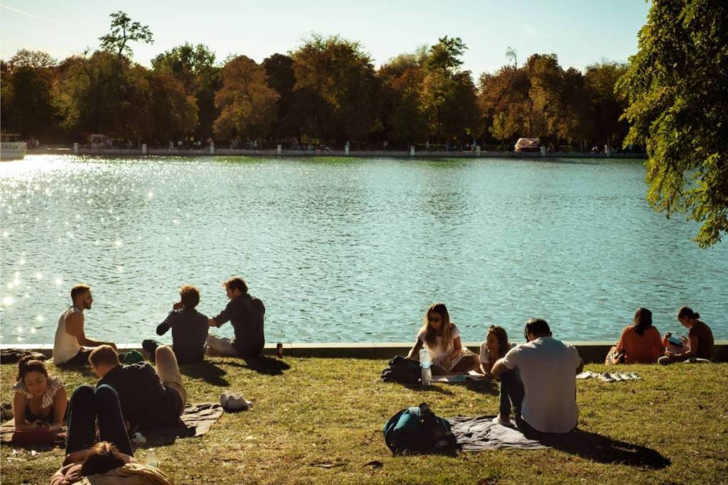 Mensen ontspannen en picknicken op een grasveld aan de waterkant in een zomers park met bomen op de achtergrond.