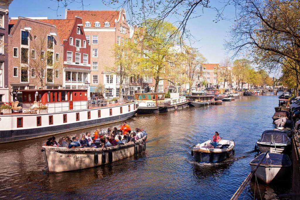 Un grand bateau ouvert bondé de monde et un petit bateau à moteur naviguent par une journée ensoleillée sur un canal d’Amsterdam, entourés de maisons historiques au bord de l’eau et de péniches.