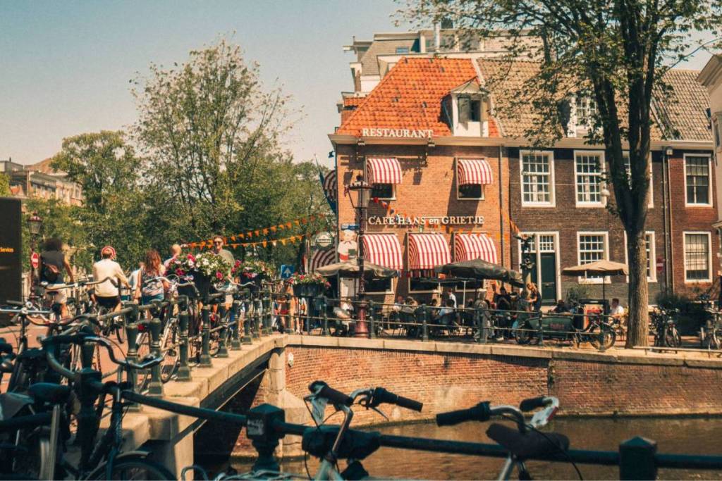 La acogedora terraza del Café Hans en Grietje junto a un canal de Ámsterdam en un día soleado, con un puente y bicicletas aparcadas en primer plano.