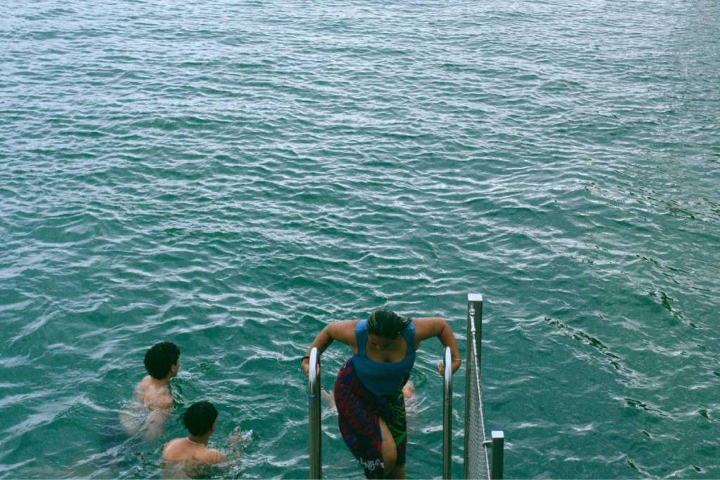 Gente que busca refrescarse en aguas cristalinas de color verde azulado, mientras una mujer sale del agua por una escalera metálica de piscina.