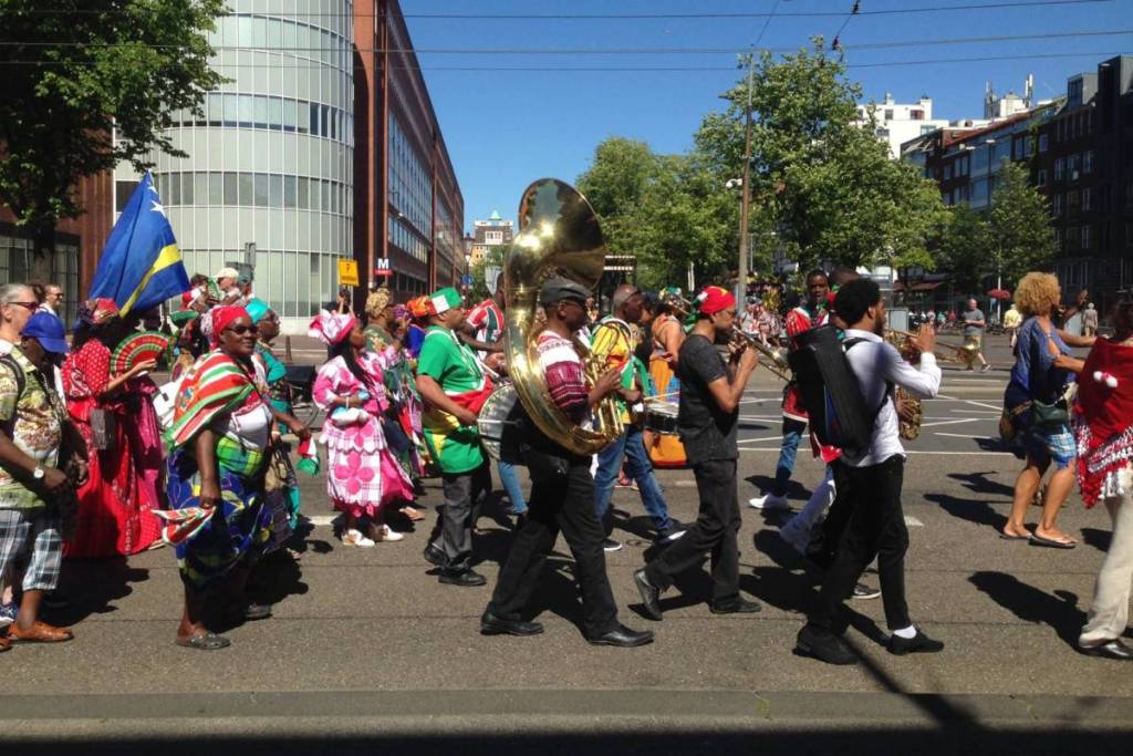 Participants in a street parade on a sunny day with a brass band. Musicians play on the street on large wind instruments such as a sousaphone and trumpets, surrounded by people marching alongside them in brightly colored clothing.
