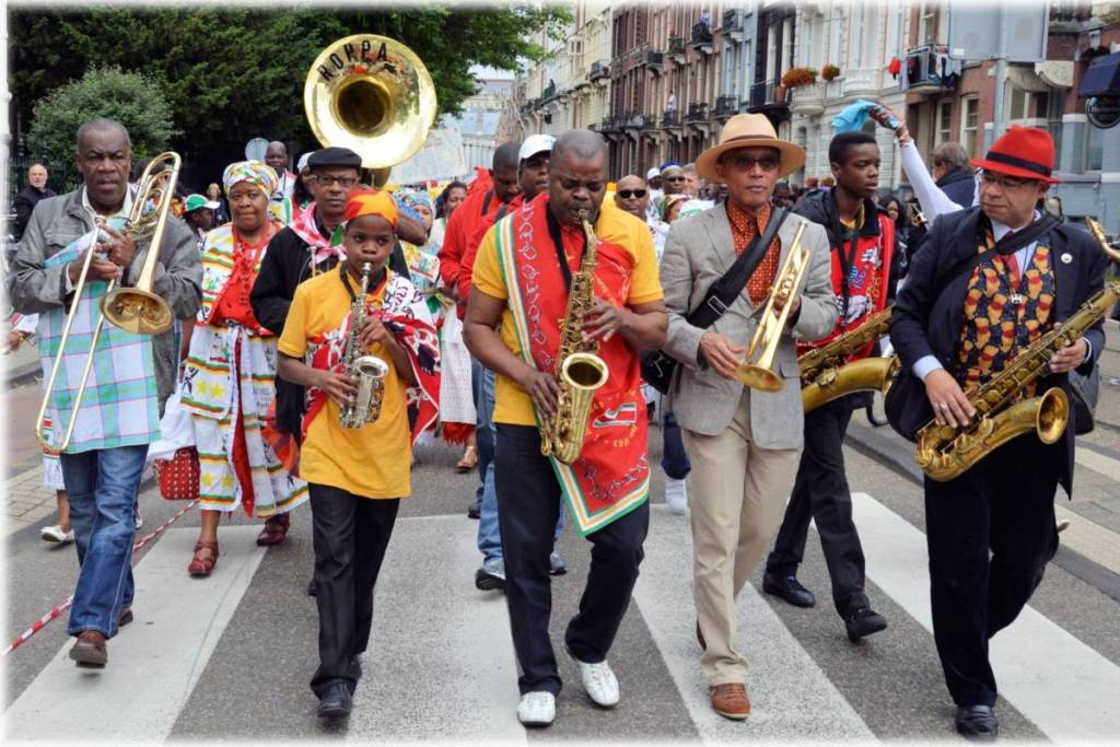 A marching band crossing a crosswalk on a city street. The musicians play various instruments, including saxophones, trumpets, and trombones.