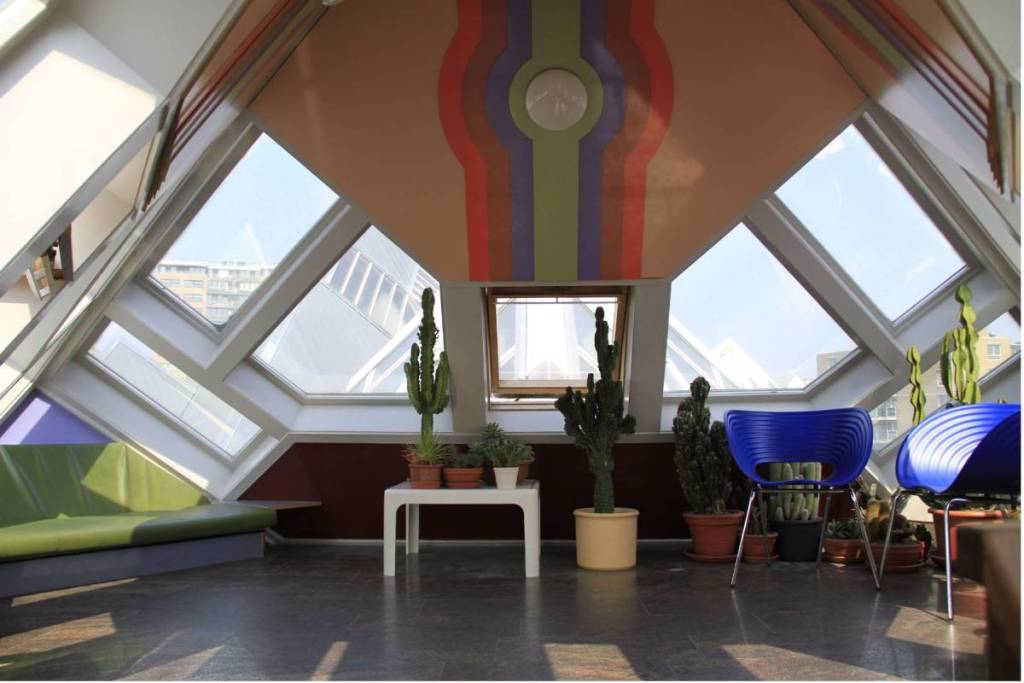 Modern interieur van een Kubuswoning in Rotterdam met schuine wanden en ramen, een groene bank, felle blauwe stoelen en cactussen onder een retro gekleurd plafond.