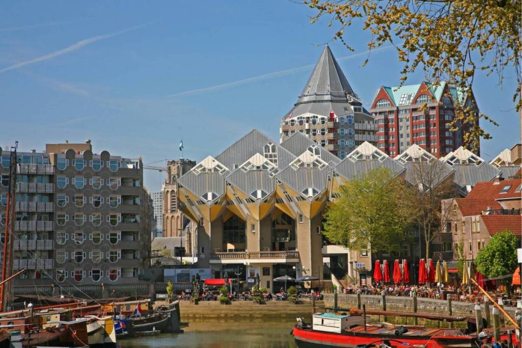 Gezicht op de gele en grijze Kubuswoningen in Rotterdam vanaf het water van de Oude Haven met historische boten op de voorgrond en de kenmerkende Blaaktoren op de achtergrond.