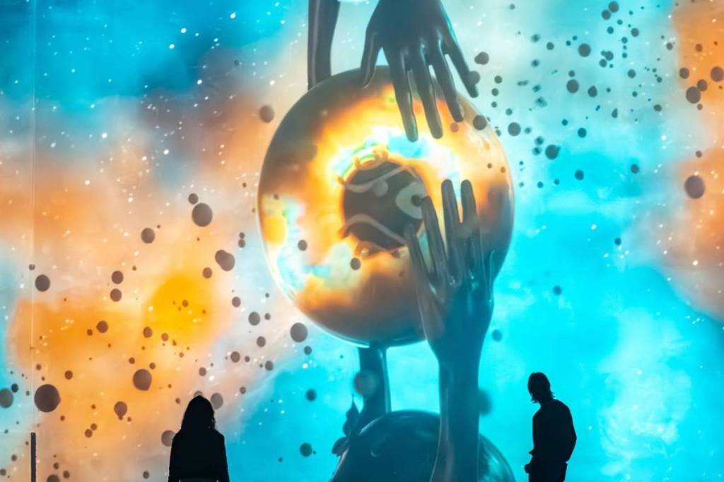 Le sagome di due visitatori alla Fabrique des Lumières che osservano una gigantesca proiezione artistica cosmica con una sfera arancione incandescente al centro, enormi mani protese e uno sfondo vivace nei toni del verde acqua e dell’arancione.