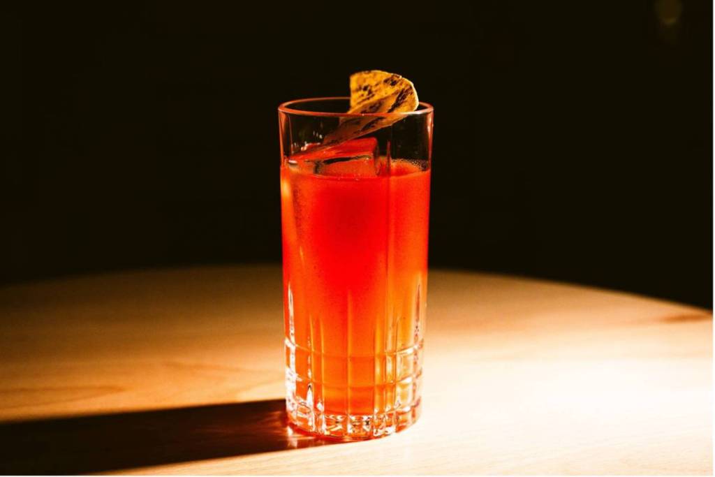 Un cocktail rouge éclatant dans un grand verre highball avec un glaçon transparent et une garniture épurée, mis en valeur de façon spectaculaire par un spot chaud sur une table en bois devant un fond sombre.