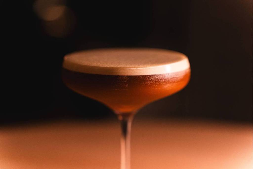 Un gros plan plein d’ambiance d’un cocktail ambré dans un élégant verre à coupe sur pied, parfaitement couronné d’une épaisse couche de mousse veloutée sous un éclairage chaleureux et intime.
