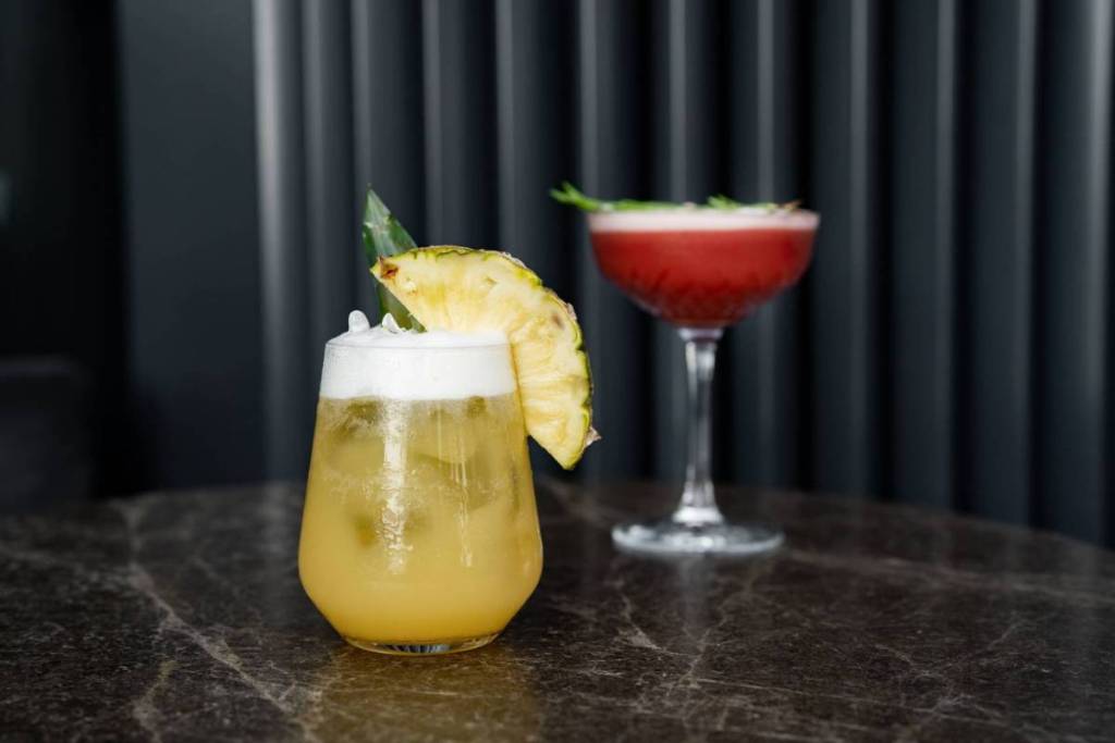Un cocktail tropical, d’un jaune doré, avec une mousse onctueuse et une généreuse tranche d’ananas frais au premier plan, derrière laquelle se profile un cocktail rouge rubis aux couleurs subtilement estompées, servi dans un élégant verre à coupe posé sur un plateau en marbre foncé.