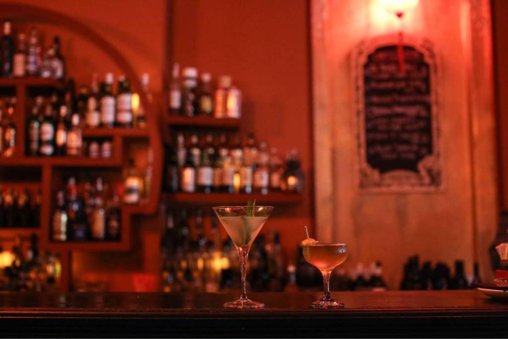 Deux cocktails raffinés – un martini agrémenté d’un brin de menthe et une coupe classique – qui brillent sur un comptoir en bois foncé, sur fond de bouteilles bien remplies éclairées d’une lumière tamisée, dans une ambiance chaleureuse aux reflets rougeoyants.