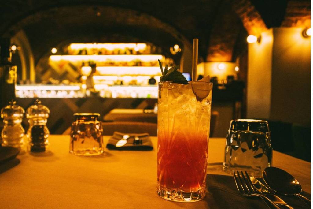 Un cocktail aux nuances dorées et rouges, servi avec une paille et de la menthe fraîche, sur une table dressée dans un restaurant plein de charme, avec un éclairage d'ambiance jaune et chaleureux et d'authentiques voûtes en briques en arrière-plan.
