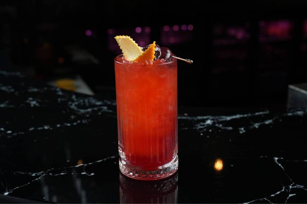 Un cocktail vif, d’un rouge rubis, rempli de glaçons dans un grand verre cannelé, garni avec raffinement d’un zeste d’orange dentelé et de cerises noires sur un pic, posé sur un comptoir en marbre noir brillant.