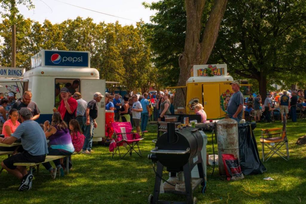 Un festival de food trucks en plein air très fréquenté, dans un parc ombragé et arboré, où les visiteurs se rassemblent autour de food trucks blancs et jaunes, avec au premier plan un grand barbecue-fumoir noir.