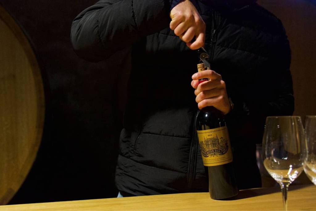 A person in a black quilted jacket uses a waiter’s knife to uncork a bottle of Alter Ego red wine at a wooden bar, with wine glasses in the foreground.