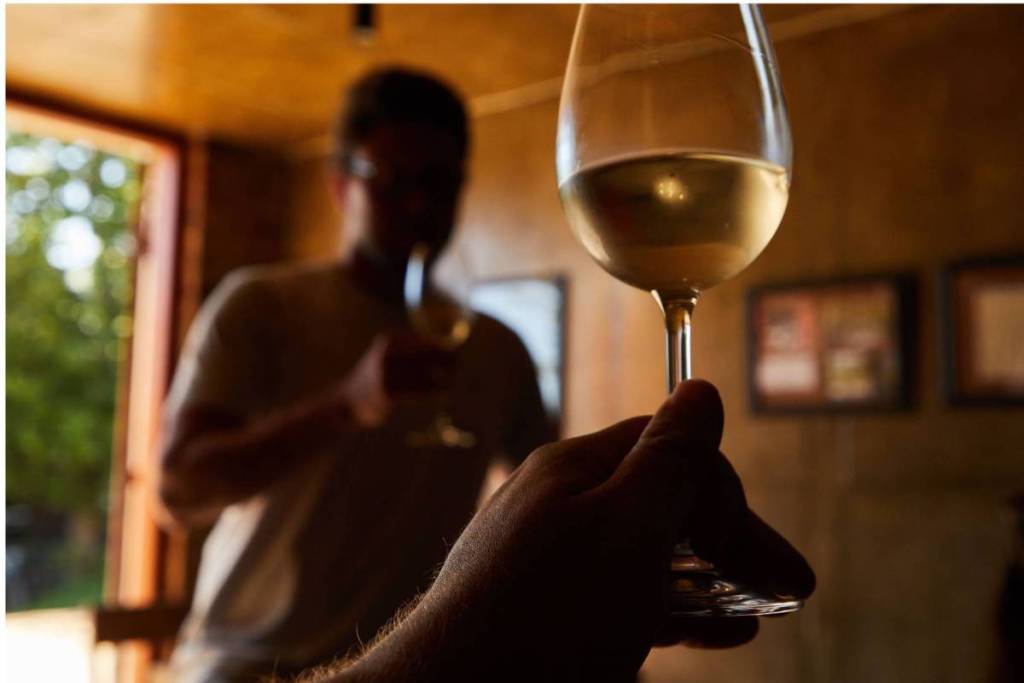 A hand holds a glass of white wine in the foreground during a tasting, with the blurred silhouette of a man sniffing his glass in the background.