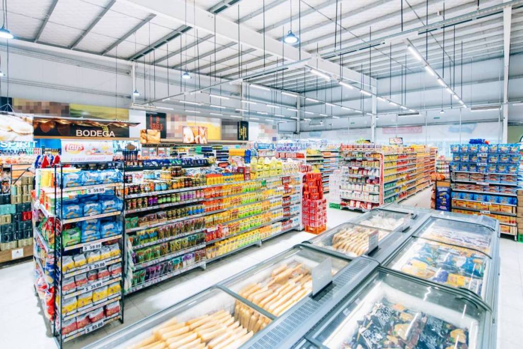 A view of a bright, well-stocked supermarket with long aisles and large freezer cases in the foreground.