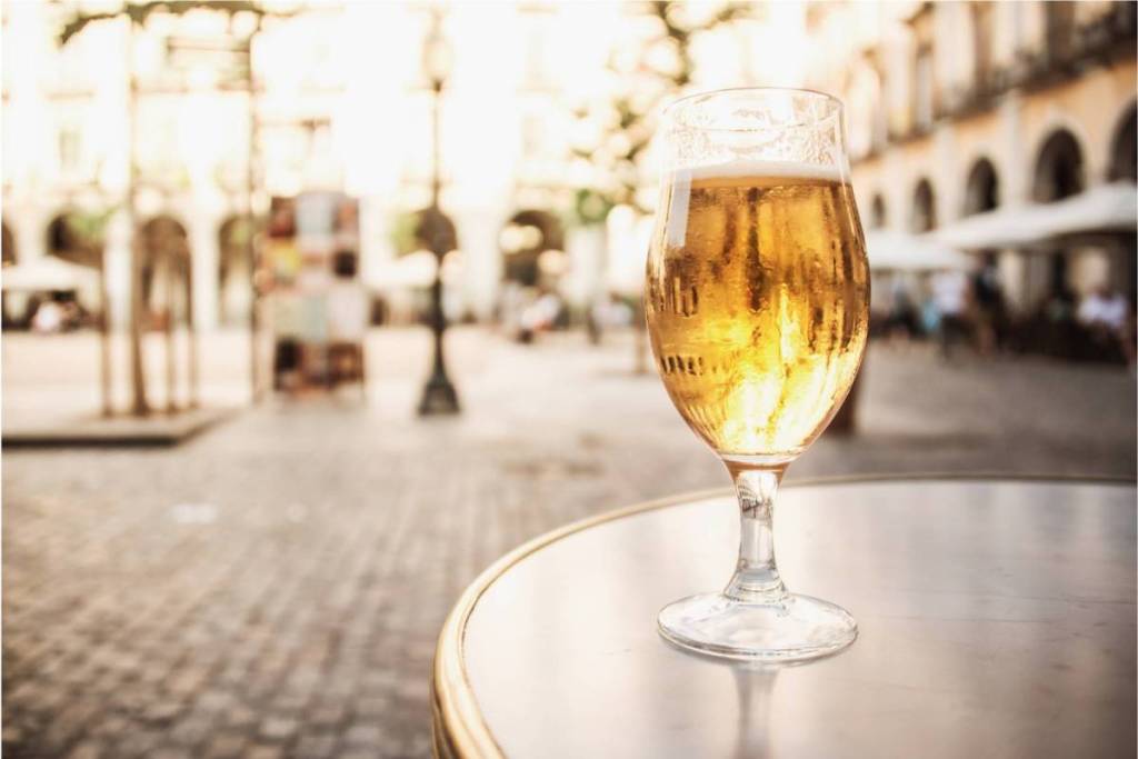 A cold glass of beer with condensation droplets on a patio table, with a sunny European city square in the background.