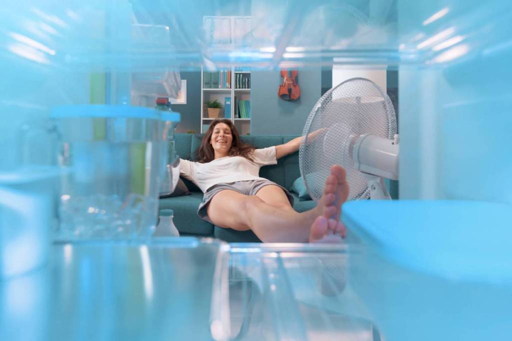 A young woman sitting relaxed on a bench in front of a spinning fan, seen from the perspective of an open refrigerator.