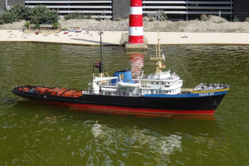 A miniature model of the tugboat Zwarte Zee sails through the waters of Madurodam, with a red-and-white lighthouse and a miniature beach in the background.