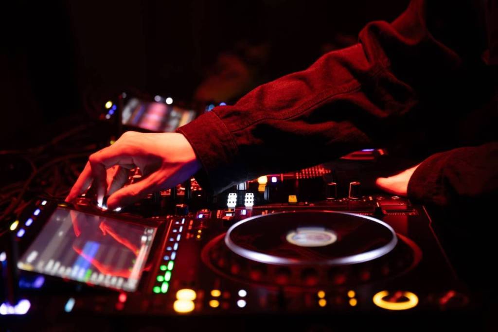 Primer plano de las manos de un DJ detrás de una mesa de mezclas iluminada en rojo, con una mano en un botón junto a la pantalla digital.