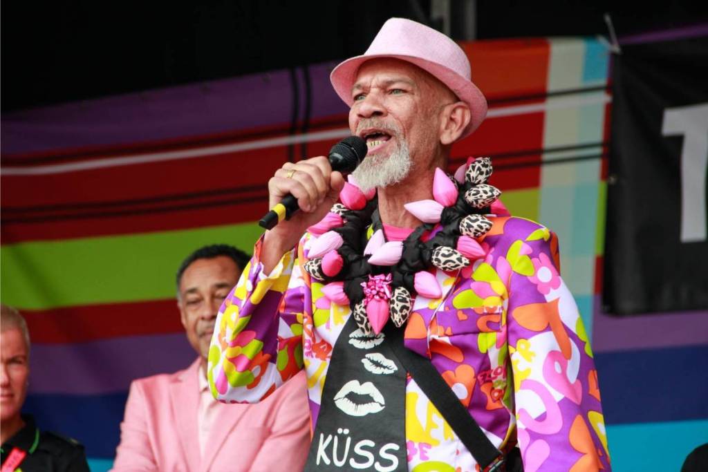 A man wearing a pink hat and a striking, colorful patterned jacket is speaking or singing into a microphone on stage. He’s wearing a pink flower garland and a black scarf.