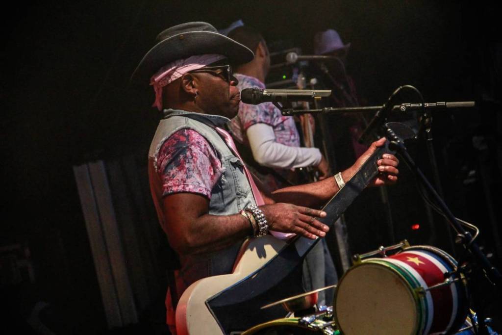 A musician plays guitar on an atmospheric, dimly lit stage. He wears a dark cowboy hat with a pink bandana underneath, sunglasses, and a denim vest. In the foreground, a drum decorated with the Surinamese flag is visible.