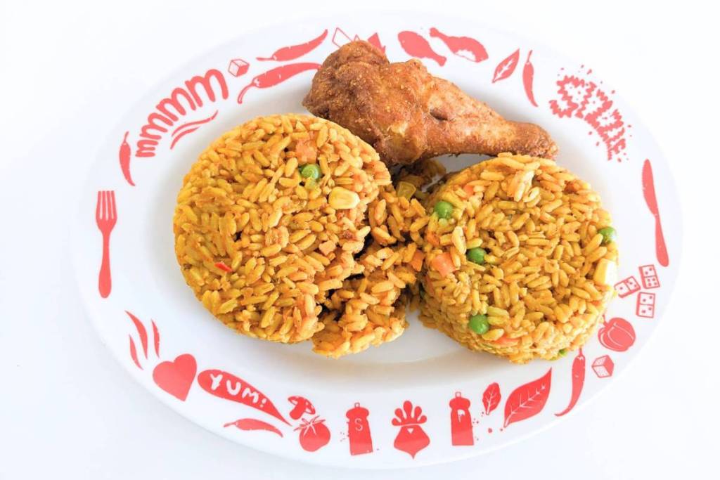 A white plate with red illustrations and text such as “YUM!” and “mmmm,” featuring two round portions of yellow spiced rice with green peas and corn, and a crispy piece of fried chicken.