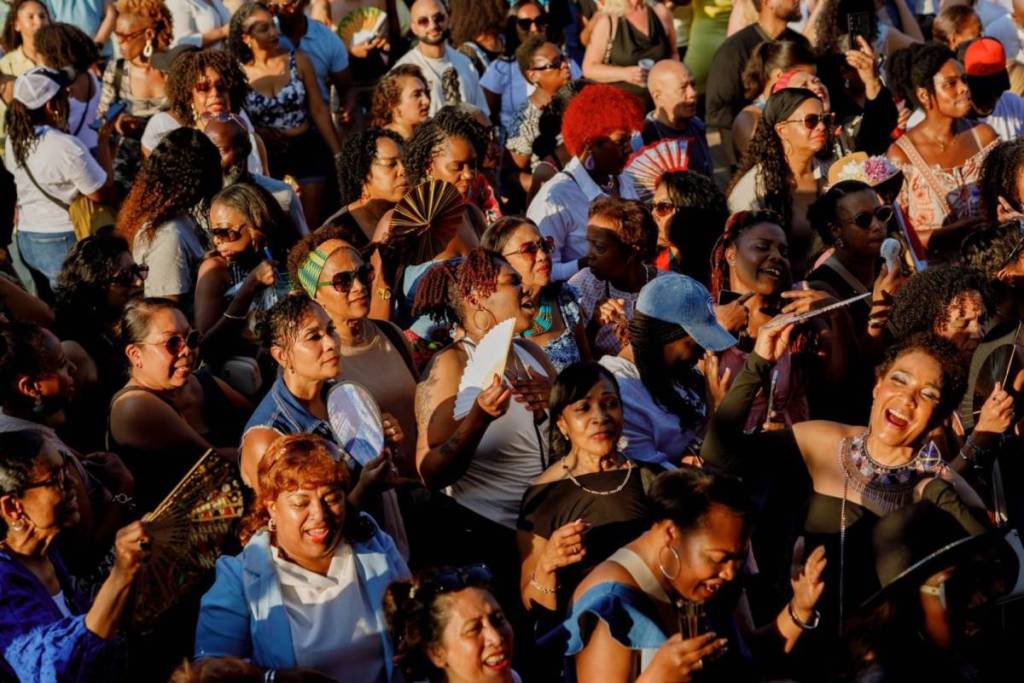 A bird’s-eye view of a large, diverse, and enthusiastic crowd enjoying the festival in the bright sunshine. Many visitors are laughing, singing along passionately, and using hand fans to cool themselves off. The crowd exudes a warm, summery, and festive atmosphere.