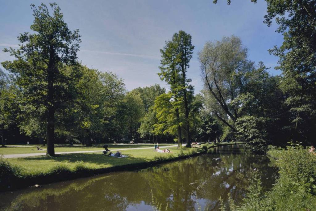 An einem sonnigen Tag entspannen sich Menschen auf einer grünen Wiese am Ufer des Vondelparks in Amsterdam und sonnen sich, während sich große Bäume im ruhigen Wasser spiegeln.