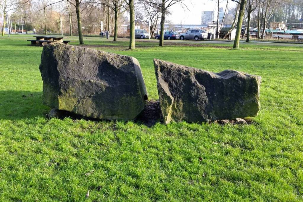 Zwei große, robuste und raue Natursteinblöcke, die direkt nebeneinander auf einer grünen Wiese im Martin-Luther-King-Park in Amsterdam liegen, im Hintergrund kahlgebliebene Bäume, ein Picknicktisch und geparkte Autos am Straßenrand.