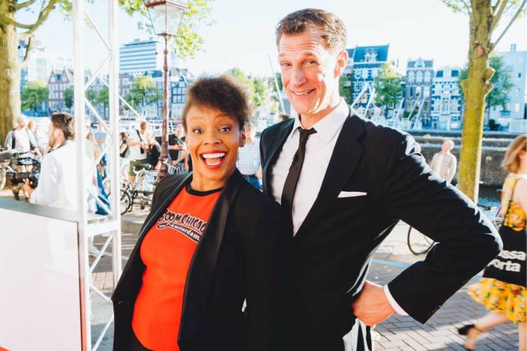 Comedienne Amber Ruffin lacht breed in een rood Boom Chicago T-shirt naast oprichter Andrew Moskos in een zwart pak, poserend langs een Amsterdamse gracht.