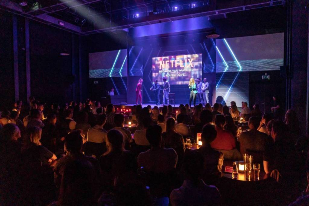 Een volle, sfeervol verlichte theaterzaal van Boom Chicago in Amsterdam kijken naar een live comedy-optreden op een verlicht podium met een groot Netflix-scherm.