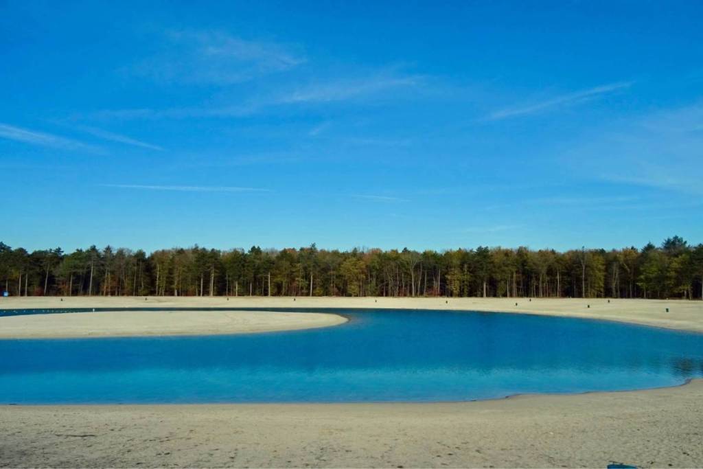 The shallow swimming pond 't Nije Hemelriek in Drenthe, with a wide sandbar and a clear blue sky above the surrounding forests.