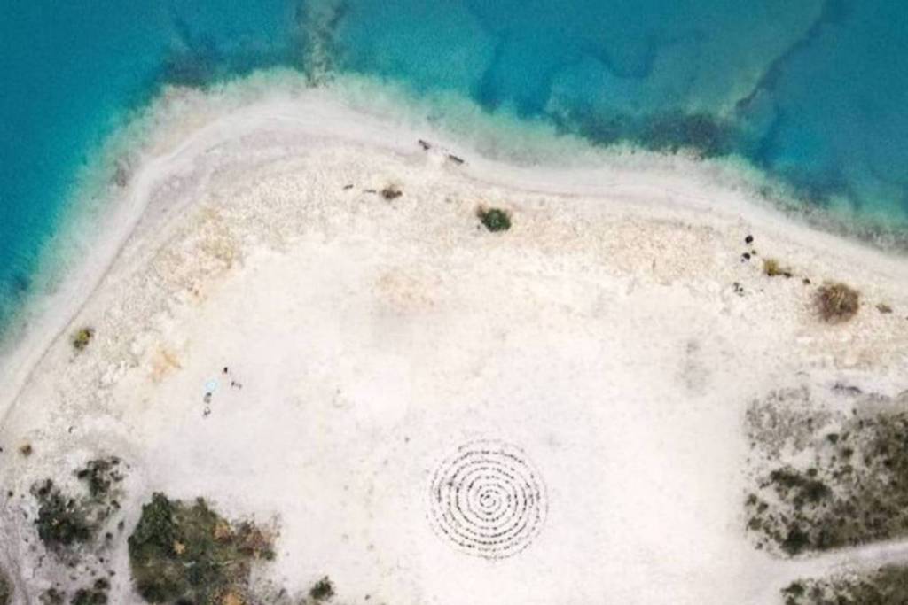 Aerial view (drone photo) of a white sandy beach with a spiral drawn in the sand, bordering the clear blue water of the recreational lake.