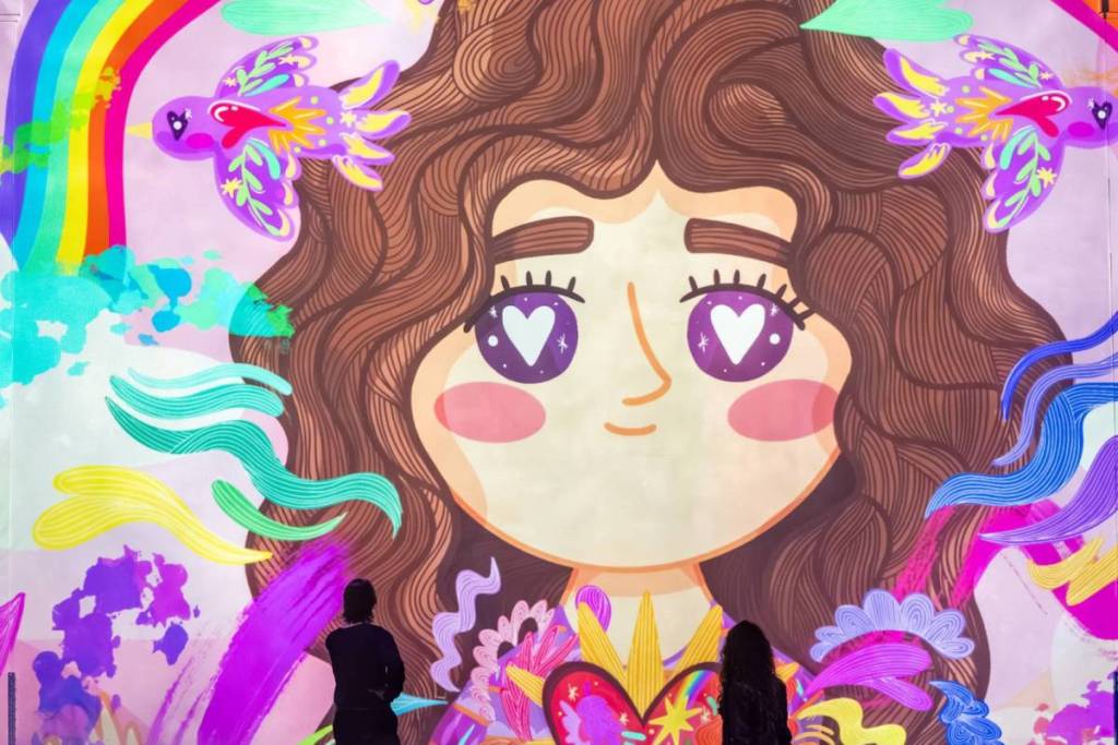 due visitatori visti da dietro mentre guardano verso l’alto una gigantesca e coloratissima proiezione murale alla Fabrique des Lumières. L’arte digitale mostra un’allegra illustrazione di una ragazzina con lunghi capelli castani ondulati e cuoricini negli occhi, circondata da un arcobaleno, uccelli viola e motivi astratti e colorati.