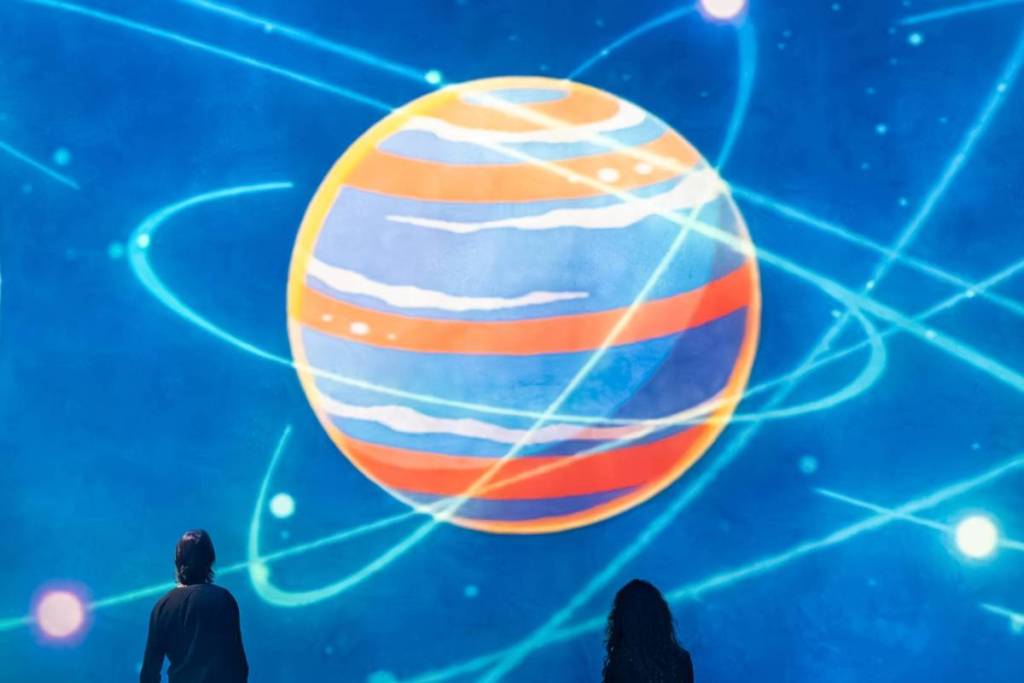 Due persone che guardano da dietro un’enorme installazione artistica cosmica di un blu intenso alla Fabrique des Lumières, con un grande pianeta a strisce colorate, anelli al neon luminosi e traiettorie stellari proiettate sulla parete.