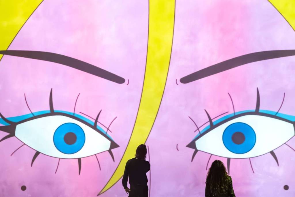 Due visitatori visti di spalle alla Fabrique des Lumières, mentre guardano una gigantesca e coloratissima proiezione murale in stile pop art che mostra un primo piano estremo di un volto disegnato con grandi occhi blu brillanti, lunghe ciglia e uno sfondo rosa e giallo.