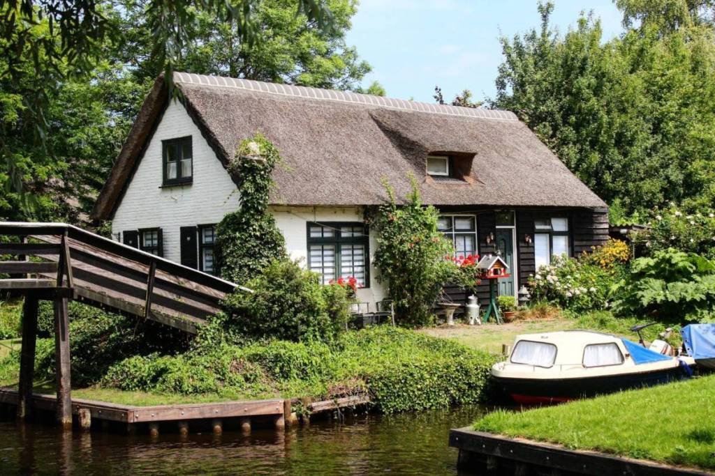 A traditional white, thatched-roof farmhouse in Giethoorn, situated on a canal with a small wooden bridge and a moored electric whisper boat.