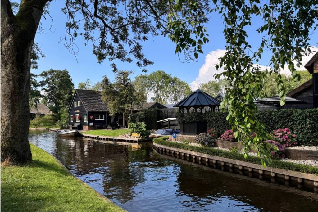 A peaceful, picturesque canal in Giethoorn surrounded by lush green trees, blooming pink hydrangeas, and characteristic black wooden houses.