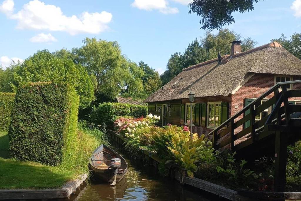 A classic wooden punt is moored in a narrow, idyllic canal next to an authentic brick farmhouse with a thatched roof and a lush flower garden.