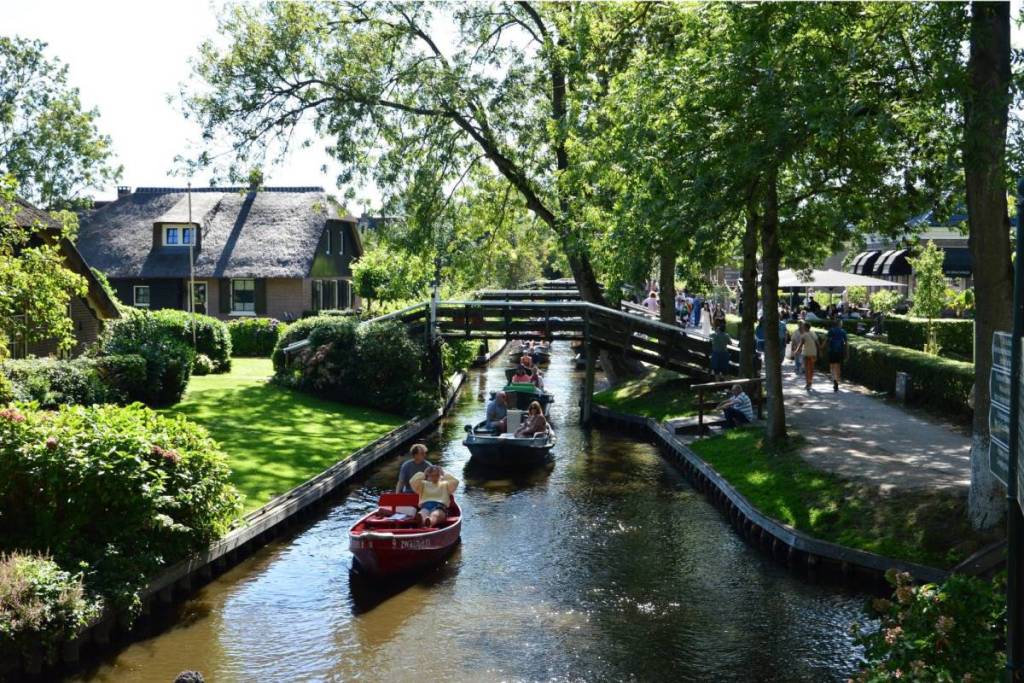 Tourists glide in electric boats along the bustling Dorpsgracht in Giethoorn, passing under a wooden arch bridge, while pedestrians stroll along the adjacent sidewalk in the sunshine.
