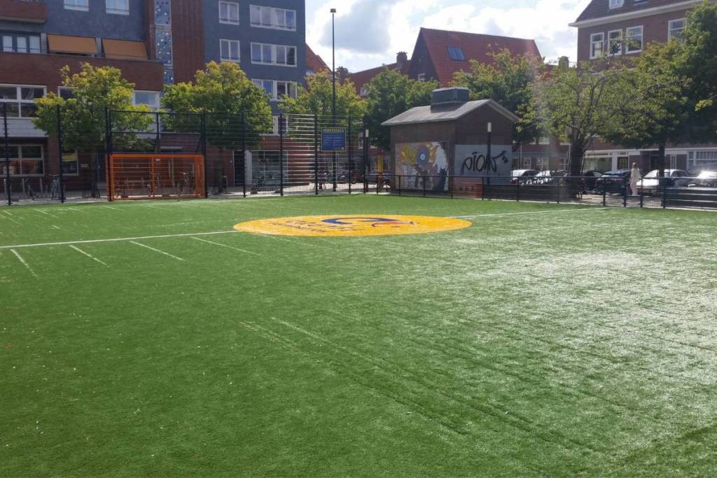 Een openbaar groen kunstgras Cruyff Court voetbalveld met het bekende oranje logo in een Amsterdamse woonwijk.